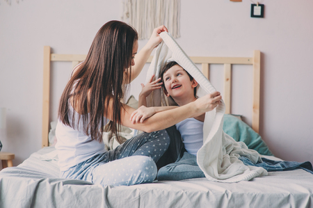 happy mother and son playing and hiding under blanket in bed in cozy weekend morning. Casual lifestyle with real familyの写真素材