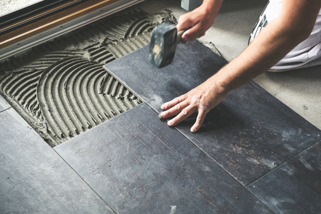 Worker placing ceramic floor tiles on adhesive surface, leveling with rubber hammer