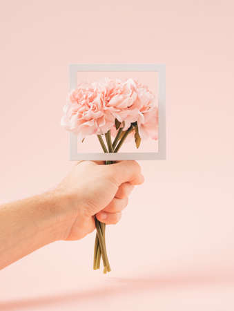 Hand holding artificial flowers bouquet with a square frame on a pastel pink background. Virtual love minimal concept.の写真素材
