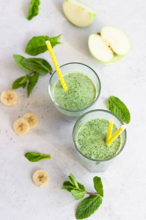 Two glasses with green spinach, banana, apple and mint smoothie with yellow straw. Light gray stone background. Healthy food concept.の写真素材
