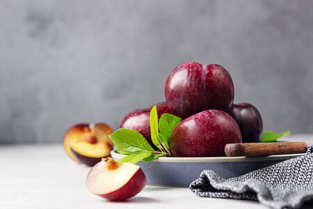 Whole purple plums and slices with leaves and knife on ceramic plate, light grey background.