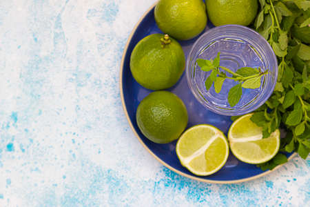 Glass with water, whole and half limes, mint on blue ceramic plate, blue background. Ingredients for...