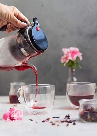 Pouring hot hibiscus tea into glass cup and pink flowers on light gray stone background. Making...