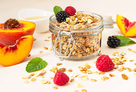 Homemade crunchy granola in glass jar, peaches, berries, honey and mint, light concrete background. Healthy vegan or vegetarian snack. Selective focus.の写真素材