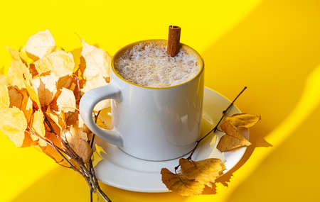 Autumn coffee or cappuccino composition. White coffee cup with milk foam, cinnamon and autumn leaves on yellow background. Fall hot drinks, cafe and bar concept. Bright sunlight and shadows.の写真素材
