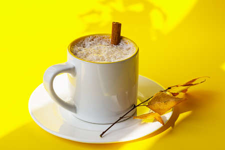 Autumn coffee or cappuccino composition. White coffee cup with milk foam, cinnamon and autumn leaves on yellow background. Fall hot drinks, cafe and bar concept. Bright sunlight and shadows.の写真素材