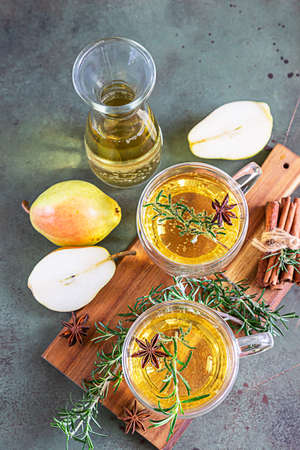 Glass mugs with hot pear or apple cider for Christmas or New Year. Traditional autumn or winter drink with rosemary, cinnamon and anise. Green concrete background. Selective focus.の写真素材
