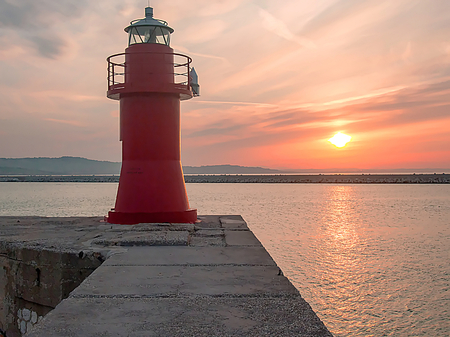The red lantern. Lighthouse isolated on the dock of Ancona's historic harbor with sunset and hot summer sky in the background and the reflection of the sun reflecting in the crisp water of the seaの写真素材