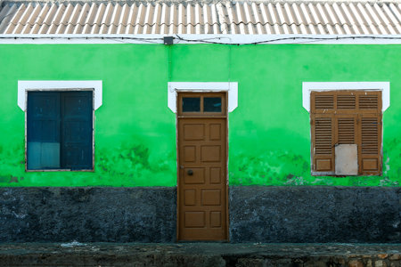 A small building with bright green walls, a central door, and windows with worn shutters. The...