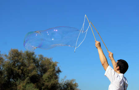 a boy who is making a big soap bubble with two sticks and a rope with a tree in the background and a...