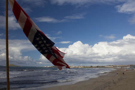 evocative image of a USA flag in the foreground waving in a sandy beach with the rough sea and...