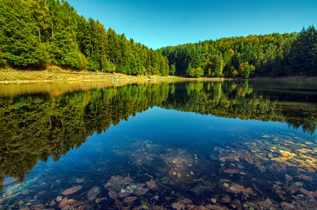 Lake Meugliano, province of Turin, Piedmont region, Italy. 8 October 2017 at 13:50. Autumn foliage colors and game of reflections on the water mirror. Green conifers around the lake. Blue sky.のeditorial素材