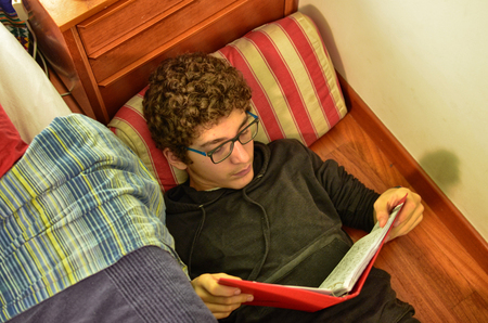 Caucasian boy with curly brown hair and blue eyeglasses, is studying on a red cover notebook.の写真素材