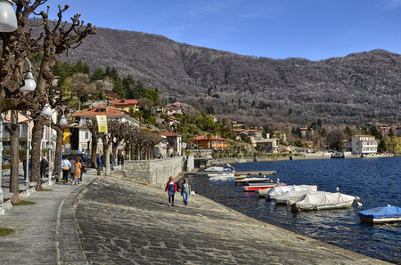 Mergozzo, Piedmont, Italy. March 2019. On the lakefront the houses with warm and bright colors face. Tourists stroll around enjoying the atmosphere, some stop on a bench. Moored boats.のeditorial素材