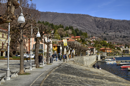 Mergozzo, Piedmont, Italy. March 2019. On the lakefront the houses with warm and bright colors face. Tourists stroll around enjoying the atmosphere, some stop on a bench. Moored boats.のeditorial素材