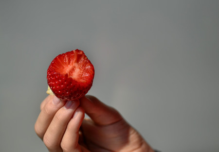 The fingers of a Caucasian boy's hand support a strawberry: it has been bitten and the glittering part with the pulp is clearly visible. Blurred background.の写真素材