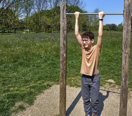 A young and friendly Caucasian boy tries his hand at pulling up the bar, raising himself with a smile. In the background the park and indistinguishable people.の写真素材