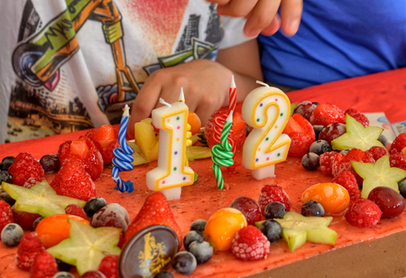 Appetizing fruit ice-cream cake decorated with strawberries, blueberries, cigliege, calamodino, kumquat, apple stars. 1 and 2 shaped candles for a 12 year birthday. Hands position and light them.の写真素材