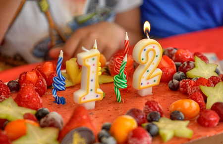Appetizing fruit ice-cream cake decorated with strawberries, blueberries, cigliege, calamodino, kumquat, apple stars. 1 and 2 shaped candles for a 12 year birthday. Hands position and light them.の写真素材