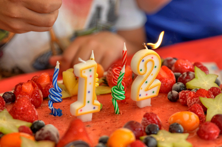 Appetizing fruit ice-cream cake decorated with strawberries, blueberries, cigliege, calamodino, kumquat, apple stars. 1 and 2 shaped candles for a 12 year birthday. Hands position and light them.の写真素材