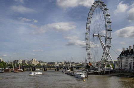 London, United Kingdom, June 2018. The London Eye photographed from the Westminster Bridge. Ships of various sizes line the Thames, gloomy clouds on blue sky.のeditorial素材