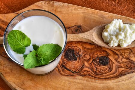 Close-up shot on a full glass of white Kefir enriched with some mint leaves. On the cutting board a wooden spoon full of Kefir ferments.の写真素材