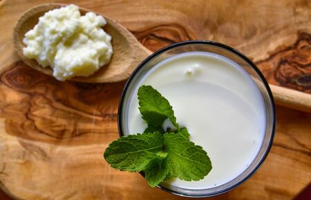 Close-up shot on a full glass of white Kefir enriched with some mint leaves. On the cutting board a wooden spoon full of Kefir ferments.の写真素材