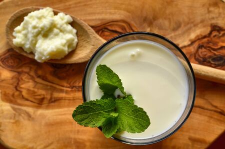 Close-up shot on a full glass of white Kefir enriched with some mint leaves. On the cutting board a wooden spoon full of Kefir ferments.の写真素材