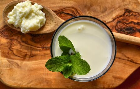 Close-up shot on a full glass of white Kefir enriched with some mint leaves. On the cutting board a wooden spoon full of Kefir ferments.の写真素材
