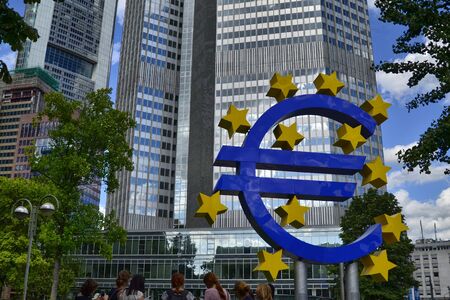 Frankfurt am Main, Germany, August 2019. Eurotower. White clouds on the blue sky. On the right is highlighted the blue euro symbol with yellow stars. People stand nearby talking.のeditorial素材
