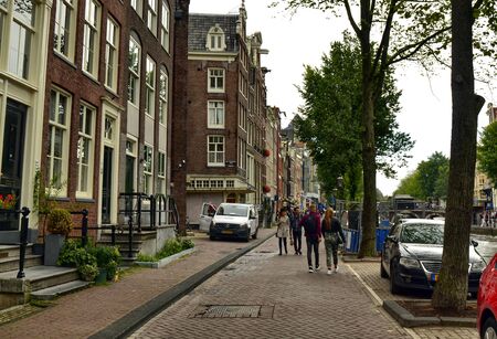 Amsterdam, Netherlands. August 2019. The streets of the historic center: on one side the typical Dutch houses, on the other the trees that line the canals. People walk.のeditorial素材