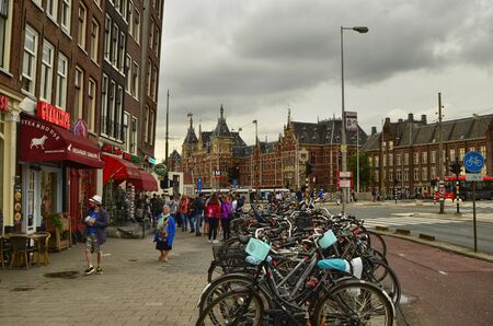 Amsterdam, Holland, August 2019. Side view of three quarters of the large central railway station. From the street a row of parked bikes leads the eye towards the station. People walk.のeditorial素材