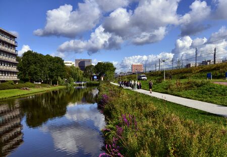 Amsterdam, Holland, August 2019. On the outskirts the canals are lined only with trees and greenery. The foliage is reflected in the water. More distant from the shore modern buildings. People walkのeditorial素材