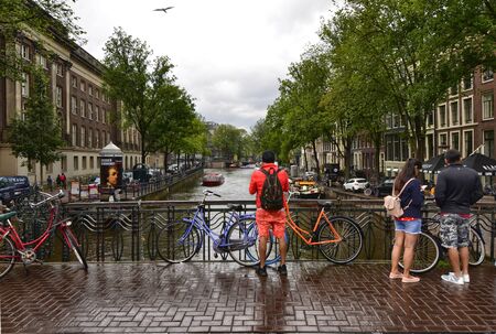 Amsterdam, Holland, August 2019. Typical view over a canal in the historic center. Rainy day. Colorful bikes parked on the railing. People cross the bridge.のeditorial素材