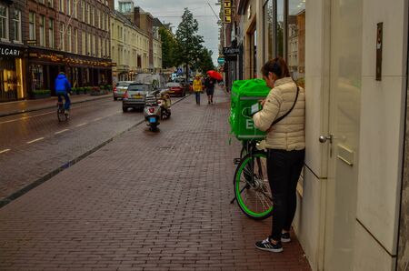 Amsterdam, Holland, August 2019. A rainy day in the shopping street. An uber eat worker consults the smartphone, next to the bike with the showy green bag to carry the food.のeditorial素材