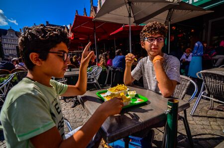 Bruges, Belgium, August 2019. A chip shop overlooking the touristy Markt square. Two Caucasian boys are enjoying the chips with mayonnaise.のeditorial素材