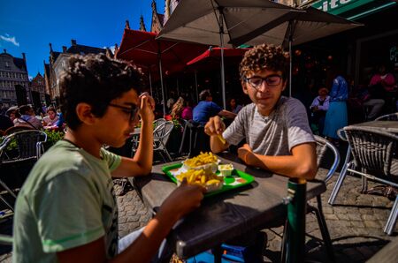 Bruges, Belgium, August 2019. A chip shop overlooking the touristy Markt square. Two Caucasian boys are enjoying the chips with mayonnaise.のeditorial素材
