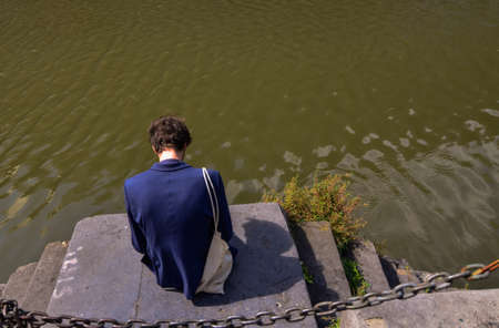 Ghent, Belgium, August 2019. Along the canals you can see students who focus on nature. A Caucasian boy, with a blue jacket and shoulder bag, is reading.のeditorial素材