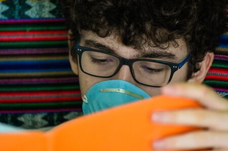Portrait of a bedridden caucasian boy wearing a blue protective mask. He is studying his...