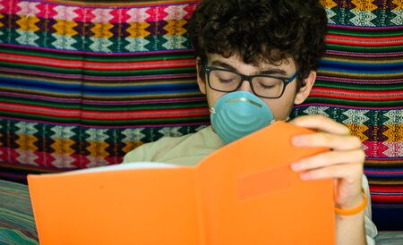 A bedridden Caucasian boy wears a blue protective mask. He is studying his orange-covered notebook....