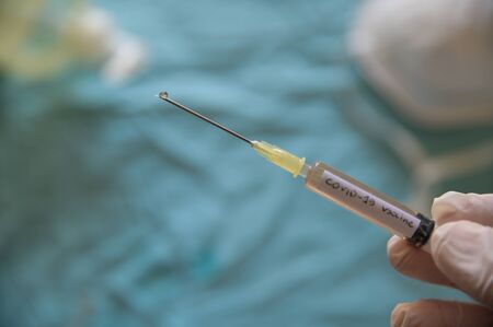 Close up shot of a syringe with the word coronavirus vaccine.The hand holding it is pressing the plunger and a drop is coming out of the needle. In the blurred background a mask and the sanitizing gelの写真素材