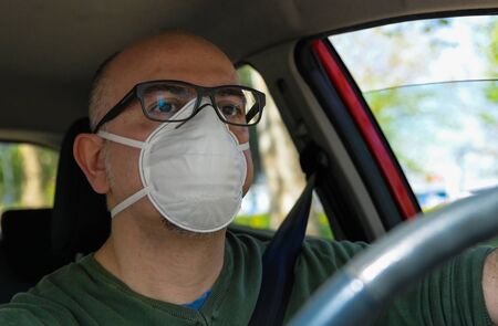 Turin, Piedmont, Italy. April 2020. Coronavirus pandemic: portrait of a Caucasian man driving the car wearing a white mask to avoid contagion. Selective focus on man and blurred background.の写真素材