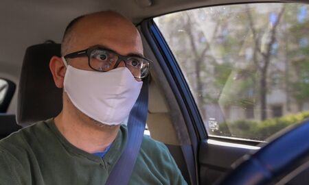 Turin, Piedmont, Italy. April 2020. Coronavirus pandemic: portrait of a Caucasian man driving the car wearing a white mask to avoid contagion. Selective focus on man and blurred background.の写真素材