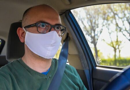 Turin, Piedmont, Italy. April 2020. Coronavirus pandemic: portrait of a Caucasian man driving the car wearing a white mask to avoid contagion. Selective focus on man and blurred background.の写真素材