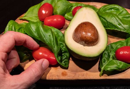 A wooden cutting board on a black background with an avocado in the center. All around, intense green basil leaves and bright red cherry tomatoes. One hand touches a cherry tomatoの写真素材