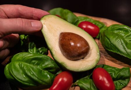 A wooden cutting board on a black background with an avocado in the center. All around, intense green basil leaves and bright red cherry tomatoes. One hand touches the avocado.の写真素材
