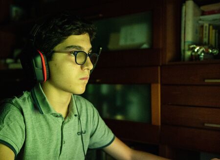 Game action during a video gaming session. The Caucasian boy with wavy black hair and glasses has his face illuminated by the changing colors of the screen during the game. Wear gaming headphones.の写真素材