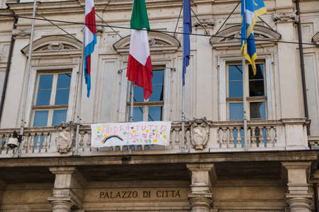 Turin, Piedmont, Italy. May 2020. Coronavirus pandemic. On the facade of the town hall, under the flags, there is a sheet with the rainbow design and the message "everything will be fine".のeditorial素材