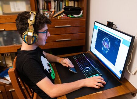 Caucasian boy with wavy brown hair and glasses wears gaming headphones. The look towards the monitor to follow the action of the video gameの写真素材
