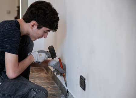 Masonry work in the apartment: a Caucasian boy is wearing work gloves and is using hammer and chisel to demolish the old baseboard.の写真素材